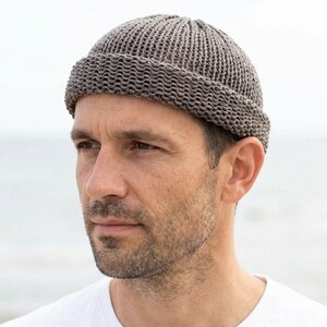 Könnte beinhalten: Eine taupefarbene Strick-Dockermütze mit einem dicken, umgeschlagenen Rand und einem strukturierten Rippenstrickmuster. Die Mütze hat eine abgerundete Krone, die eng am Kopf anliegt und eine gleichmäßige, gewebte Garnstruktur aufweist.