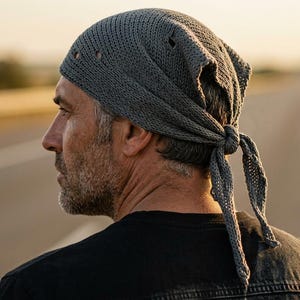 Könnte beinhalten: Graue Strickkopfbedeckung mit Knoten hinten. Die Person, die sie trägt, hat einen Bart und dunkle Kleidung. Der Hintergrund zeigt eine Straße und einen Sonnenuntergang mit Unschärfe.