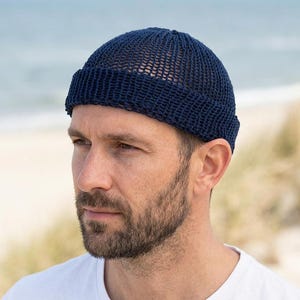 May include: A navy blue knit beanie with a rolled brim. The hat is made of a breathable, open-weave material. The person in the image is wearing a white t-shirt and has a beard. The background shows a beach and ocean.