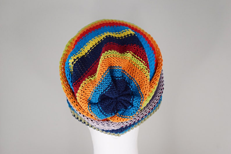 May include: A knit beanie featuring horizontal stripes in vibrant colors including navy blue, bright orange, lime green, turquoise, and light purple. The hat has a gathered crown design with a solid navy blue center point and a textured knit pattern.