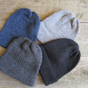 May include: Four knit beanies in solid colors arranged in a cluster. The collection includes one dark blue beanie, one light gray beanie, one charcoal gray beanie, and one solid black beanie. Each hat features a ribbed cuff and a simple knit texture.