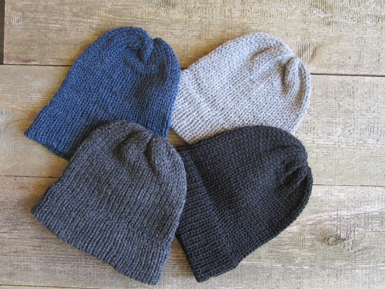 May include: Four knit beanies in solid colors arranged in a cluster. The collection includes one dark blue beanie, one light gray beanie, one charcoal gray beanie, and one solid black beanie. Each hat features a ribbed cuff and a simple knit texture.