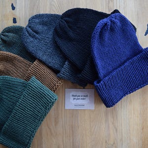 K&ouml;nnte beinhalten: Eine Sammlung von f&uuml;nf gestrickten M&uuml;tzen in Unifarben, darunter Dunkelgr&uuml;n, Braun, Anthrazit, Schwarz und K&ouml;nigsblau, jeweils mit einem breiten umgeschlagenen Rand. In der Mitte liegt eine kleine rechteckige Karte mit dem Text Thank you so much for your order.