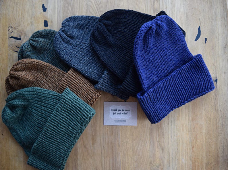 K&ouml;nnte beinhalten: Eine Sammlung von f&uuml;nf gestrickten M&uuml;tzen in Unifarben, darunter Dunkelgr&uuml;n, Braun, Anthrazit, Schwarz und K&ouml;nigsblau, jeweils mit einem breiten umgeschlagenen Rand. In der Mitte liegt eine kleine rechteckige Karte mit dem Text Thank you so much for your order.