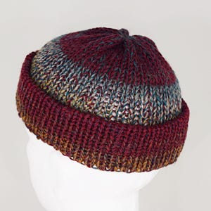 Pu&ograve; includere: Un berretto con risvolto caratterizzato da una trama a maglia grossa. Il design presenta una sfumatura di colore che passa dal bordeaux intenso sulla corona e sul bordo a un mix variegato di toni grigi, blu e dorati nella parte centrale.