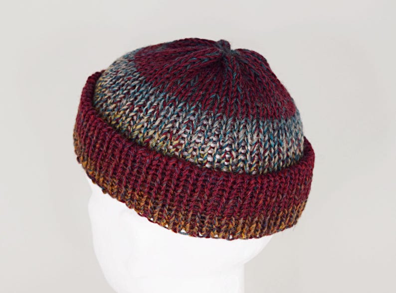 Pu&ograve; includere: Un berretto con risvolto caratterizzato da una trama a maglia grossa. Il design presenta una sfumatura di colore che passa dal bordeaux intenso sulla corona e sul bordo a un mix variegato di toni grigi, blu e dorati nella parte centrale.
