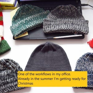 Pu&ograve; includere: Una collezione di cinque berretti lavorati a maglia in vari colori tra cui verde, grigio, nero e blu, insieme a un articolo a righe rosse e verdi con due pon pon. Il testo recita: One of the workflows in my office. Already in the summer I'm getting ready for Christmas.