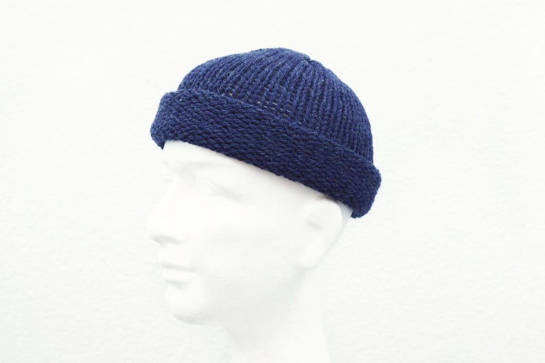 May include: A dark navy blue knit beanie cap featuring a thick, folded brim and a ribbed texture throughout the crown. The hat is constructed with a dense, woven yarn pattern and sits securely on a plain, featureless white mannequin head.