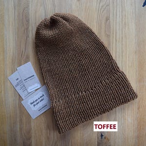 K&ouml;nnte beinhalten: Eine einfarbige braune Strickm&uuml;tze mit gerippter Textur und umgeschlagenem Rand. Neben der M&uuml;tze liegen zwei kleine rechteckige Karten mit aufgedrucktem Text, darunter der Satz Thank you so much for your order. Das Wort TOFFEE ist in Rot gedruckt.