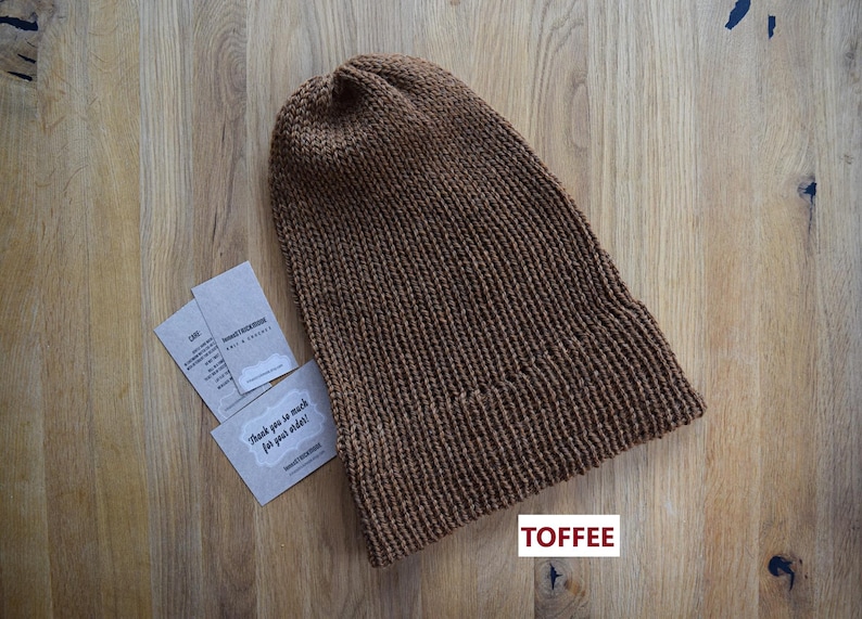 K&ouml;nnte beinhalten: Eine einfarbige braune Strickm&uuml;tze mit gerippter Textur und umgeschlagenem Rand. Neben der M&uuml;tze liegen zwei kleine rechteckige Karten mit aufgedrucktem Text, darunter der Satz Thank you so much for your order. Das Wort TOFFEE ist in Rot gedruckt.