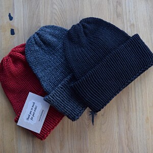 K&ouml;nnte beinhalten: Drei Strickm&uuml;tzen in Rot, Anthrazit und Schwarz, jeweils mit umgeschlagenem B&uuml;ndchen. In der roten M&uuml;tze steckt eine kleine rechteckige wei&szlig;e Karte mit dem aufgedruckten Text Thank you so much for your order! in einer dunklen Schreibschrift.
