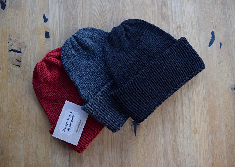 K&ouml;nnte beinhalten: Drei Strickm&uuml;tzen in Rot, Anthrazit und Schwarz, jeweils mit umgeschlagenem B&uuml;ndchen. In der roten M&uuml;tze steckt eine kleine rechteckige wei&szlig;e Karte mit dem aufgedruckten Text Thank you so much for your order! in einer dunklen Schreibschrift.