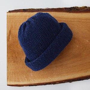 May include: A solid navy blue knit beanie hat featuring a thick, folded cuff at the base. The hat is constructed from a textured yarn with a visible ribbed stitch pattern, creating a rounded crown shape that sits compactly on the surface.