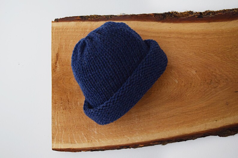 May include: A solid navy blue knit beanie hat featuring a thick, folded cuff at the base. The hat is constructed from a textured yarn with a visible ribbed stitch pattern, creating a rounded crown shape that sits compactly on the surface.