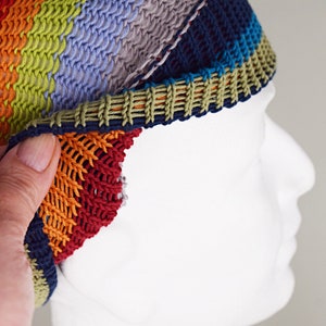 May include: A multi-colored knit beanie featuring horizontal stripes in shades of navy blue, red, orange, lime green, and light purple. The hat has a textured, open-stitch construction with a solid navy blue border and a contrasting olive green trim.