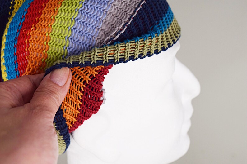 May include: A multi-colored knit beanie featuring horizontal stripes in shades of navy blue, red, orange, lime green, and light purple. The hat has a textured, open-stitch construction with a solid navy blue border and a contrasting olive green trim.