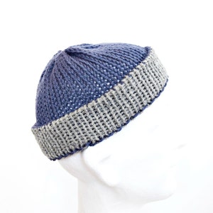 Könnte beinhalten: Eine Strickmütze mit einer dunkelblauen Krone in vertikaler Rippenstruktur und einem kontrastierenden, umgeschlagenen hellgrauen Rand. Die Mütze ist in einem groben Strickmuster gefertigt und zeigt eine deutliche Farbtrennung zwischen dem blauen Oberteil und dem grauen Umschlag.