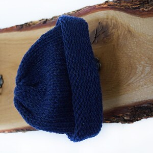 May include: A dark navy blue knit beanie with a thick folded cuff. The beanie is constructed from a textured yarn with a visible stitch pattern and is displayed hanging on a metal hook attached to a natural wood plank with a live edge.
