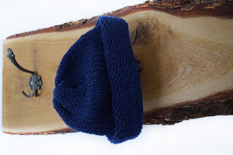 May include: A dark navy blue knit beanie with a thick folded cuff. The beanie is constructed from a textured yarn with a visible stitch pattern and is displayed hanging on a metal hook attached to a natural wood plank with a live edge.