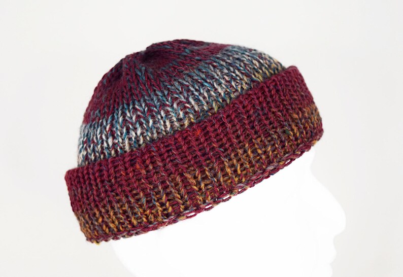 Pu&ograve; includere: Un berretto in maglia con bordo risvoltato e un motivo a filato variegato. Il cappello presenta una ricca tavolozza di colori bordeaux profondo, rosso scuro e sottili sfumature di blu e oro nella struttura in lana lavorata a mano.