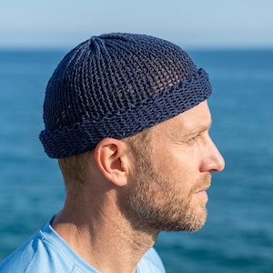 May include: A navy blue knit beanie with a folded brim. The hat is made of a textured, open-weave material. The beanie is worn by a person with a beard and short hair, with a blue ocean in the background.
