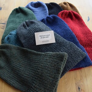 K&ouml;nnte beinhalten: Eine Sammlung von gestrickten M&uuml;tzen in verschiedenen Farben, darunter Dunkelgr&uuml;n, Anthrazit, Blau, Schwarz, Braun und Rot. Auf der anthrazitfarbenen M&uuml;tze liegt eine kleine rechteckige beigefarbene Karte mit dem Text Thank you so much for your order!