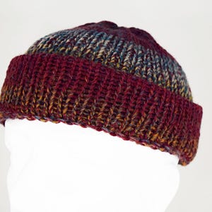 Pu&ograve; includere: Un berretto con risvolto caratterizzato da una trama a maglia grossa. Il berretto presenta una tavolozza di colori variegati composta da bordeaux profondo, rosso scuro e sfumature di marrone terroso e blu tenue nel filato, creando un aspetto multicolore.