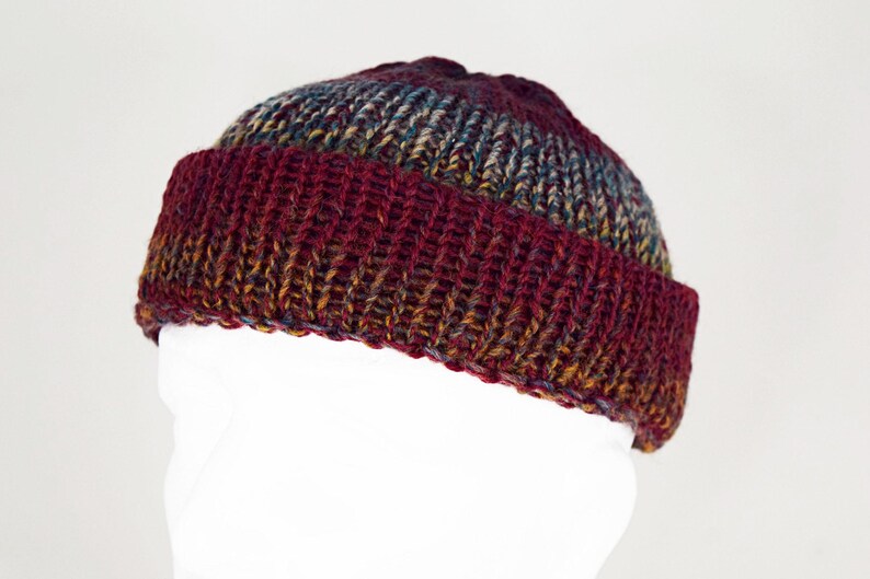 Pu&ograve; includere: Un berretto con risvolto caratterizzato da una trama a maglia grossa. Il berretto presenta una tavolozza di colori variegati composta da bordeaux profondo, rosso scuro e sfumature di marrone terroso e blu tenue nel filato, creando un aspetto multicolore.