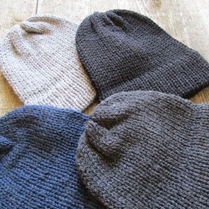 May include: Four knitted beanies in solid colors including light gray, dark charcoal gray, navy blue, and a medium gray. Each hat features a simple ribbed knit texture with a rounded crown and a folded brim, crafted from thick yarn material.