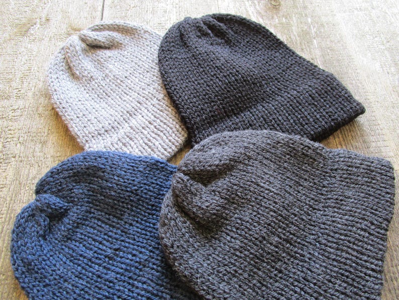 May include: Four knitted beanies in solid colors including light gray, dark charcoal gray, navy blue, and a medium gray. Each hat features a simple ribbed knit texture with a rounded crown and a folded brim, crafted from thick yarn material.