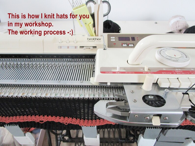 May include: A close-up of a Brother knitting machine in a workshop. The machine is cream-colored with a metal bed, currently knitting a black and coral hat. Text on the machine reads "This is how I knit hats for you in my workshop. The working process -:)".
