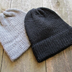 May include: Two knit beanies, one in light gray and one in solid black. Both hats feature a thick, ribbed texture with a folded cuff at the base and a rounded crown. The construction shows a consistent yarn stitch pattern throughout each hat.