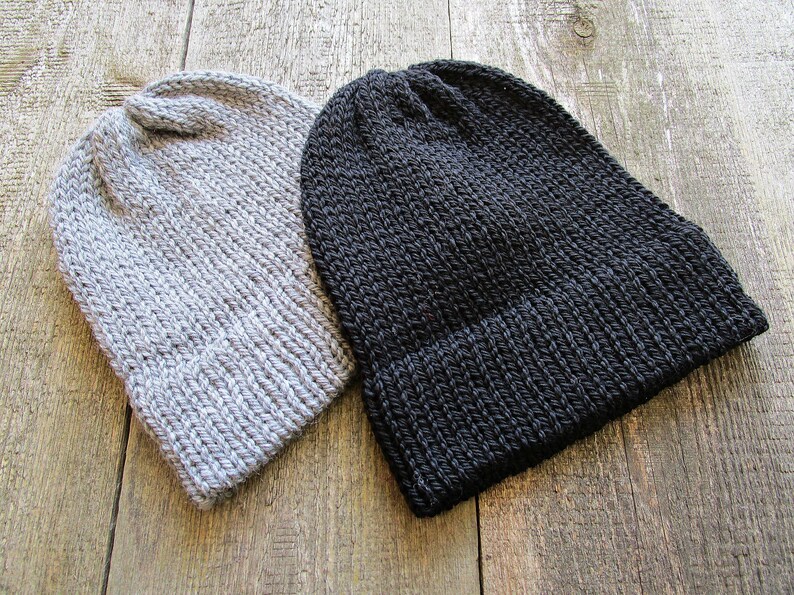 May include: Two knit beanies, one in light gray and one in solid black. Both hats feature a thick, ribbed texture with a folded cuff at the base and a rounded crown. The construction shows a consistent yarn stitch pattern throughout each hat.
