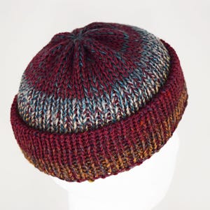 Pu&ograve; includere: Un berretto lavorato a maglia a mano con bordo risvoltato e una corona con un motivo a diminuzioni circolari. Il filato presenta una tavolozza di colori variegati in bordeaux intenso, blu tenue e tocchi di marrone dorato, creando un aspetto testurizzato.