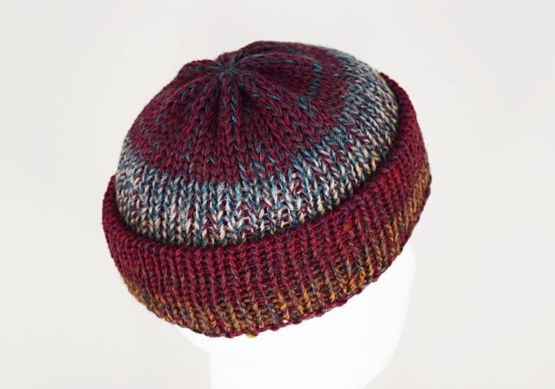 Pu&ograve; includere: Un berretto lavorato a maglia a mano con bordo risvoltato e una corona con un motivo a diminuzioni circolari. Il filato presenta una tavolozza di colori variegati in bordeaux intenso, blu tenue e tocchi di marrone dorato, creando un aspetto testurizzato.