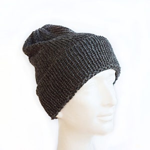 K&ouml;nnte beinhalten: Eine dunkel anthrazitfarbene Strickm&uuml;tze mit geripptem Strukturmuster. Die M&uuml;tze hat eine leicht lockere Passform und wird auf einem wei&szlig;en Schaufensterpuppenkopf pr&auml;sentiert, wobei die vertikalen Strickmaschen und der umgeschlagene Saum sichtbar sind.