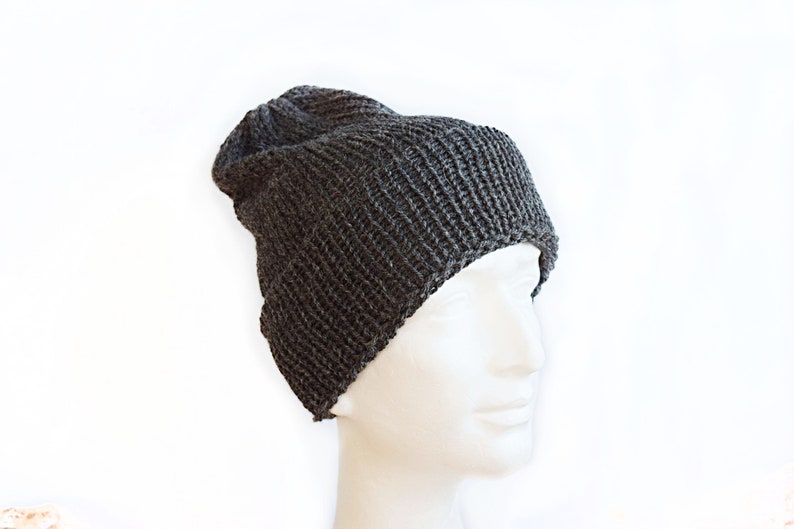 K&ouml;nnte beinhalten: Eine dunkel anthrazitfarbene Strickm&uuml;tze mit geripptem Strukturmuster. Die M&uuml;tze hat eine leicht lockere Passform und wird auf einem wei&szlig;en Schaufensterpuppenkopf pr&auml;sentiert, wobei die vertikalen Strickmaschen und der umgeschlagene Saum sichtbar sind.