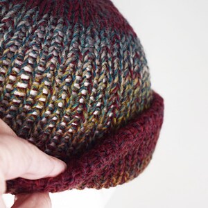 Pu&ograve; includere: Un berretto lavorato a maglia a mano con un motivo di filato variegato nei toni del bordeaux profondo, verde bosco e giallo senape. Il cappello presenta un bordo spesso ripiegato in tinta bordeaux scuro e una lavorazione a coste.