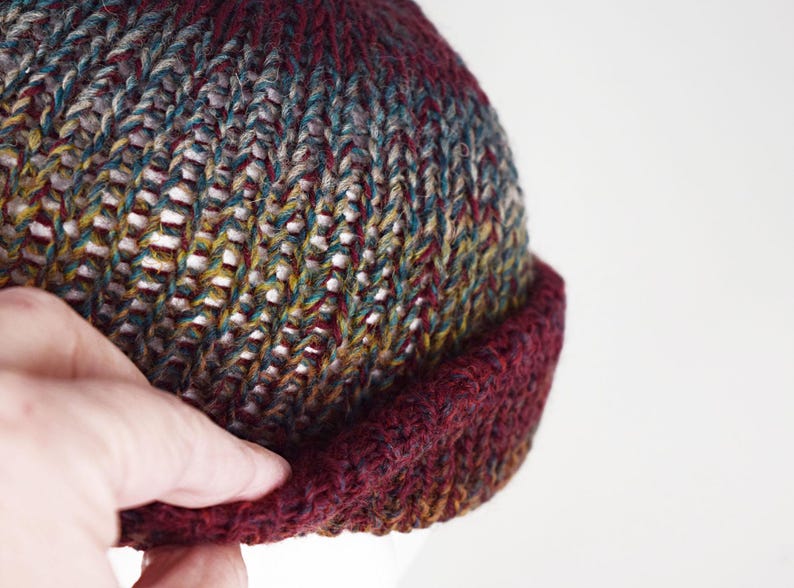 Pu&ograve; includere: Un berretto lavorato a maglia a mano con un motivo di filato variegato nei toni del bordeaux profondo, verde bosco e giallo senape. Il cappello presenta un bordo spesso ripiegato in tinta bordeaux scuro e una lavorazione a coste.