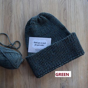 K&ouml;nnte beinhalten: Eine dunkelgr&uuml;ne Strickm&uuml;tze mit breitem Umschlag, begleitet von einem passenden Wollkn&auml;uel und einer kleinen rechteckigen Karte mit dem Text "Thank you so much for your order!" und "KNITSTITCH" in schwarzer Schrift gedruckt.
