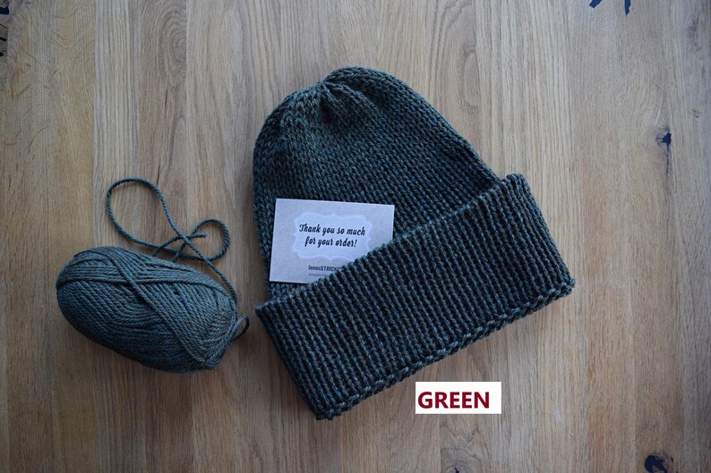 K&ouml;nnte beinhalten: Eine dunkelgr&uuml;ne Strickm&uuml;tze mit breitem Umschlag, begleitet von einem passenden Wollkn&auml;uel und einer kleinen rechteckigen Karte mit dem Text "Thank you so much for your order!" und "KNITSTITCH" in schwarzer Schrift gedruckt.