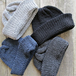 May include: Four knit beanies in light gray, black, navy blue, and dark charcoal gray. Each hat features a thick, folded ribbed cuff and a textured knit pattern throughout the crown, showcasing a classic winter headwear design in four colors.