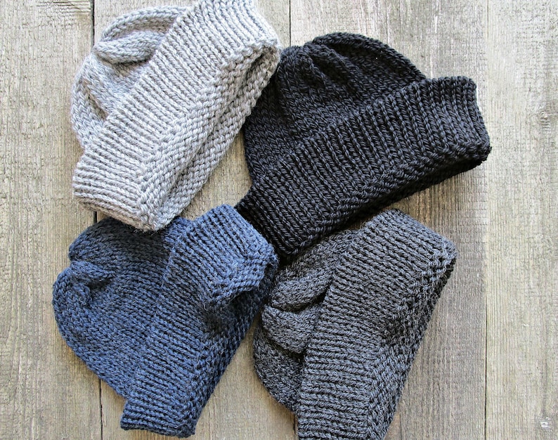 May include: Four knit beanies in light gray, black, navy blue, and dark charcoal gray. Each hat features a thick, folded ribbed cuff and a textured knit pattern throughout the crown, showcasing a classic winter headwear design in four colors.
