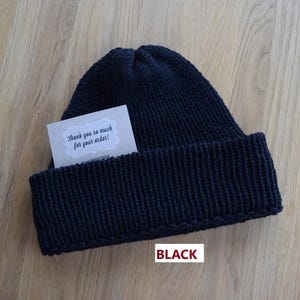 K&ouml;nnte beinhalten: Eine einfarbige schwarze Strickm&uuml;tze mit breitem Umschlag. In den Umschlag ist eine kleine rechteckige Karte mit gewelltem Rand gesteckt, die den gedruckten Text "Thank you so much for your order!" enth&auml;lt. Das Wort "BLACK" ist in Rot unter der M&uuml;tze gedruckt.