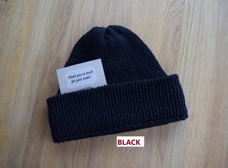 K&ouml;nnte beinhalten: Eine einfarbige schwarze Strickm&uuml;tze mit breitem Umschlag. In den Umschlag ist eine kleine rechteckige Karte mit gewelltem Rand gesteckt, die den gedruckten Text "Thank you so much for your order!" enth&auml;lt. Das Wort "BLACK" ist in Rot unter der M&uuml;tze gedruckt.