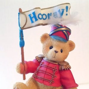 May include: A teddy bear figurine wearing a red and gold uniform with a blue and white hat. The bear is holding a blue and white banner that says "Hooray!" The bear is standing on a stone base.