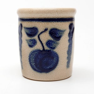 May include: A small, cream-colored ceramic jar with a blue and white floral design. The jar has a blue apple with leaves on the front.