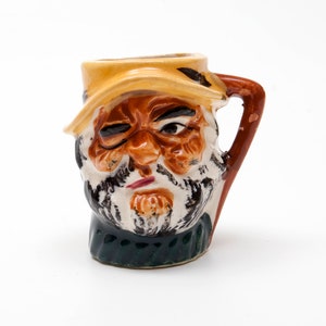 May include: A ceramic mug shaped like a man's head with a brown beard and a yellow hat. The mug has a brown handle and a green and brown design on the bottom.