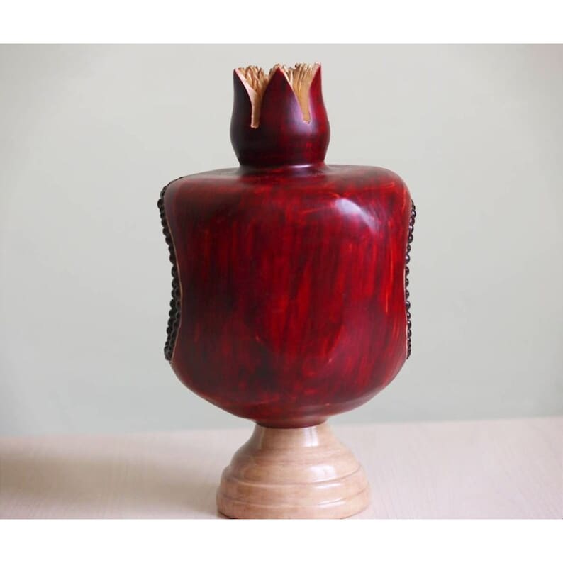May include: A wooden pomegranate sculpture with a red finish and a natural wood base. The pomegranate is carved with details and has a textured surface.