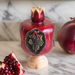 May include: A red wooden pomegranate ornament with a carved design of grapes and leaves. The ornament is inscribed with the word "Armenia" on the side.